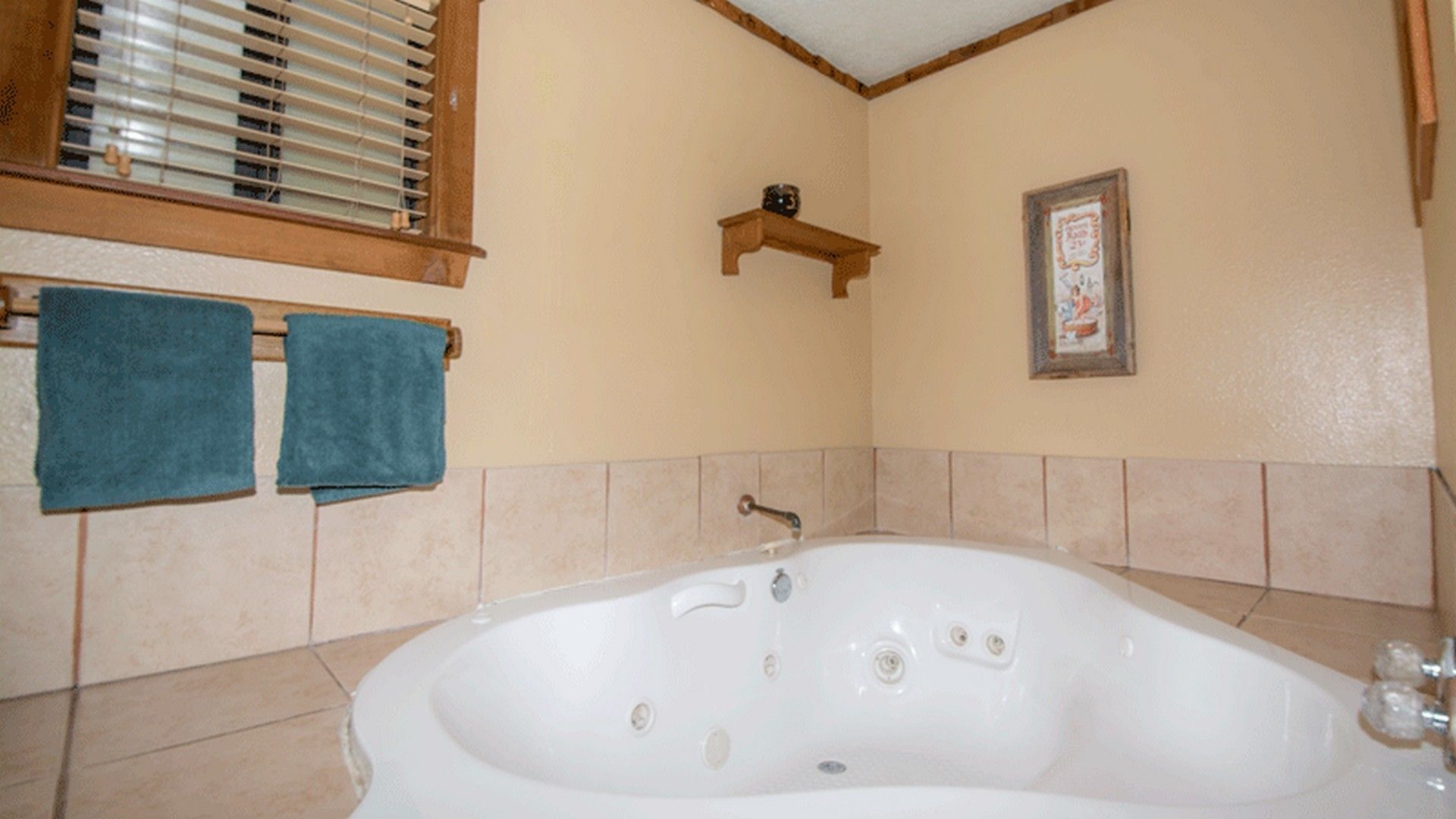 A bathroom with a bathtub, two towels, a window with blinds, a shelf with a small item, and a framed picture on the wall.