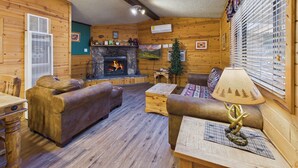 A cozy log cabin interior with a stone fireplace, wooden furniture, and a large window with blinds. - Whispering Pine Cabins (Ruidoso)