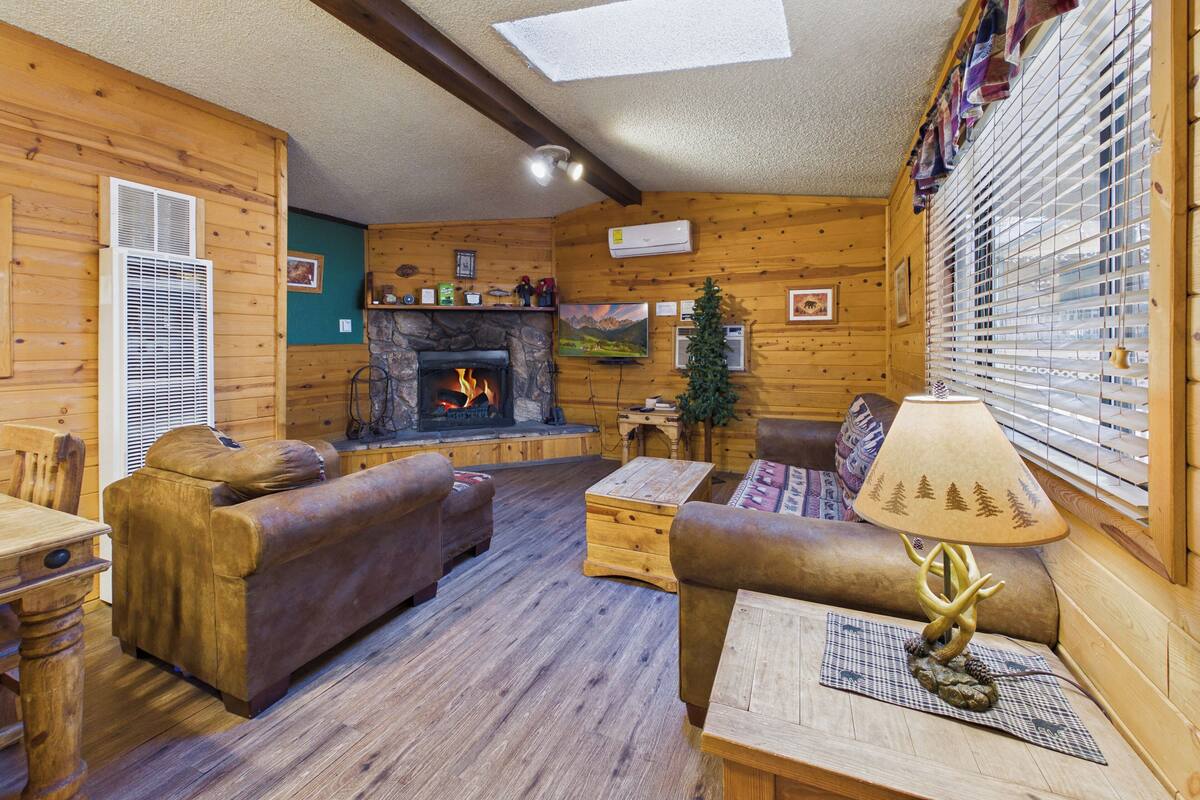 L’intérieur d’un chalet confortable, avec des murs en rondins, une cheminée en pierre, des meubles en bois et une grande fenêtre équipée de stores.