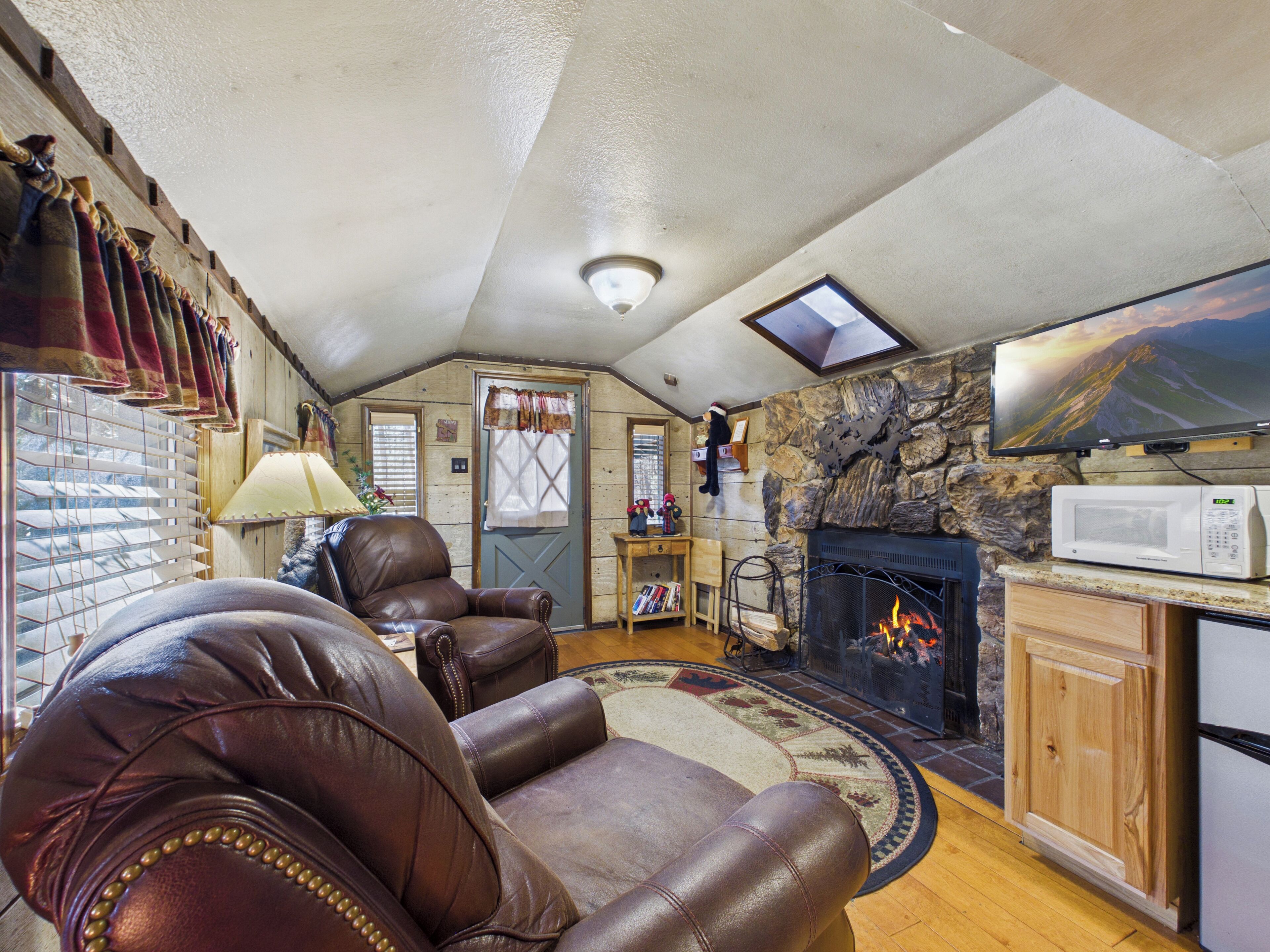 A cozy living room with leather chairs, a fireplace, a stone wall, a television, and a wooden cabinet.