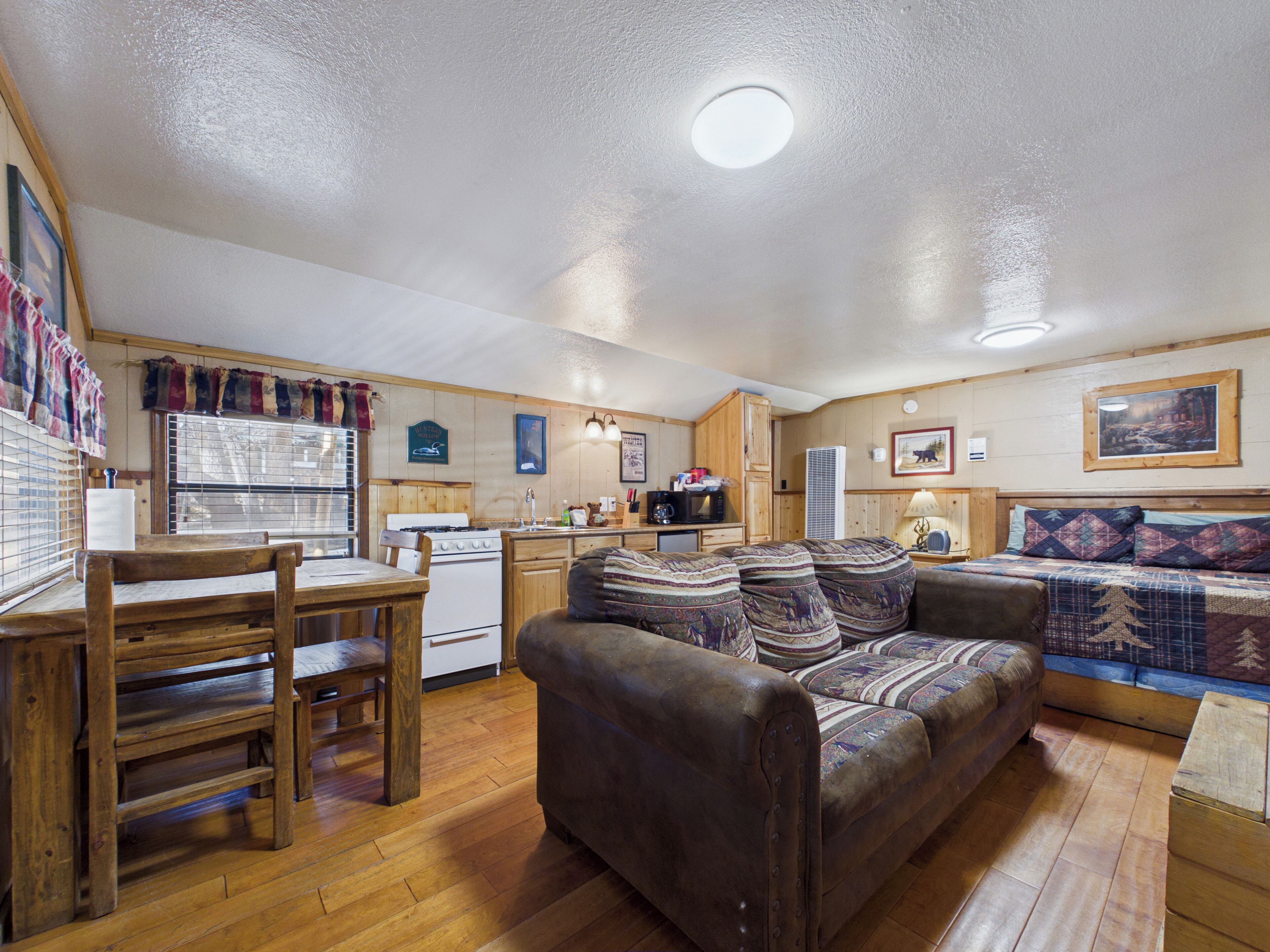 A cozy cabin interior with a wooden dining table, a sofa, a kitchen area, and a bed with patterned bedding.