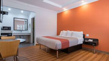 Une chambre d’hôtel avec un lit, une table de chevet et un petit bureau avec une chaise. La salle de bain est équipée d’un lavabo et d’un miroir. La chambre a un plancher en bois et un plafond avec des spots encastrés.