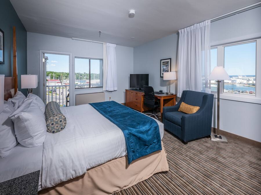 A hotel room with a bed, desk, chair, and large windows offering a view of the city.