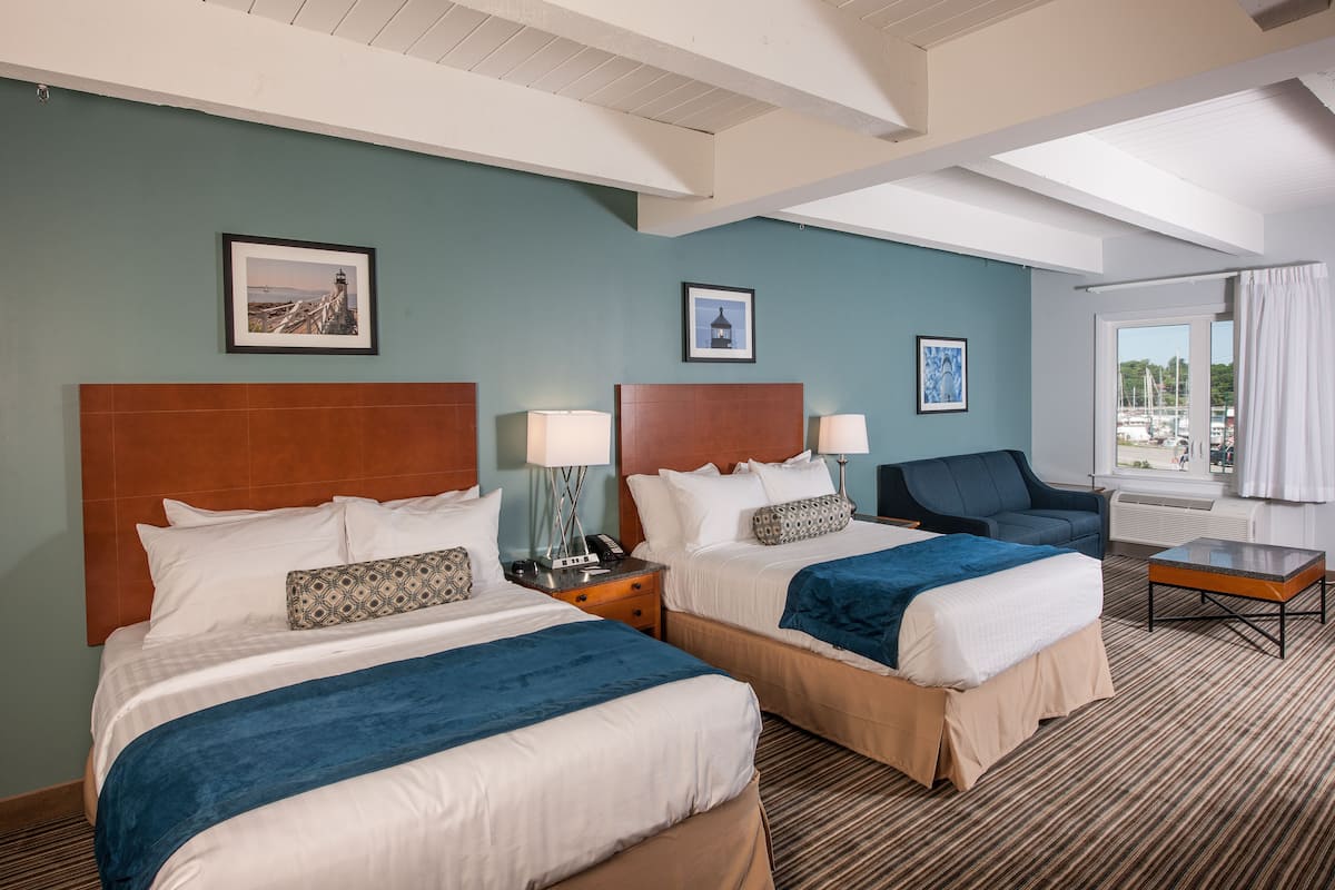 A hotel room with two beds, a sofa, a table, and a window with a view of boats.