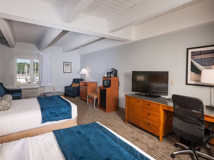 A hotel room with two beds, a desk, a chair, a television, and a window with a view of boats.