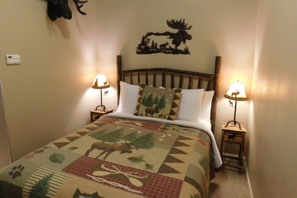 Un llit amb un estoretat estampat, dues làmpades laterals, un capçal de fusta i decoracions a la paret que inclouen la silueta d'un alce i banya.