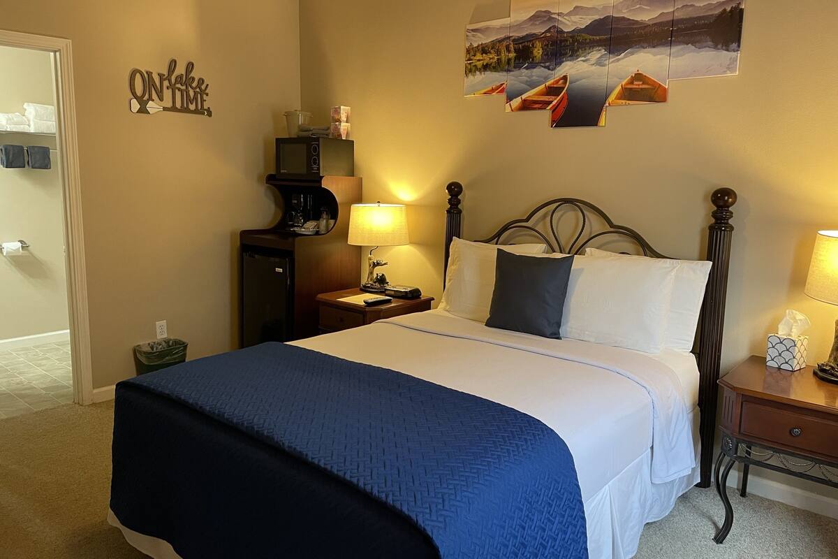 Habitació d'hotel amb llit, una taula de nit, una làmpada, taula petita, paperera, nevera, prestatgeries fixes amb productes de tocador i una decoració mural de vaixells a un llac.