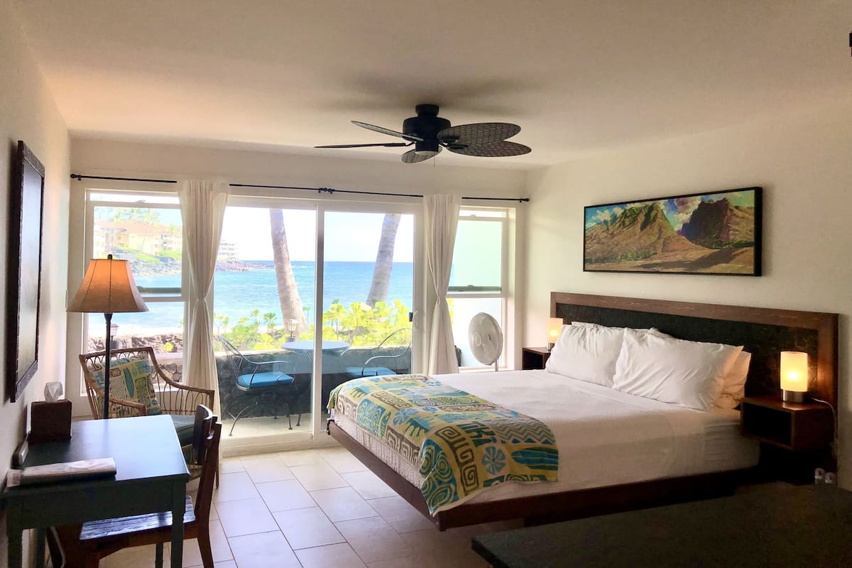 A hotel room with a bed, a desk, a chair, a fan, and a view of the ocean.