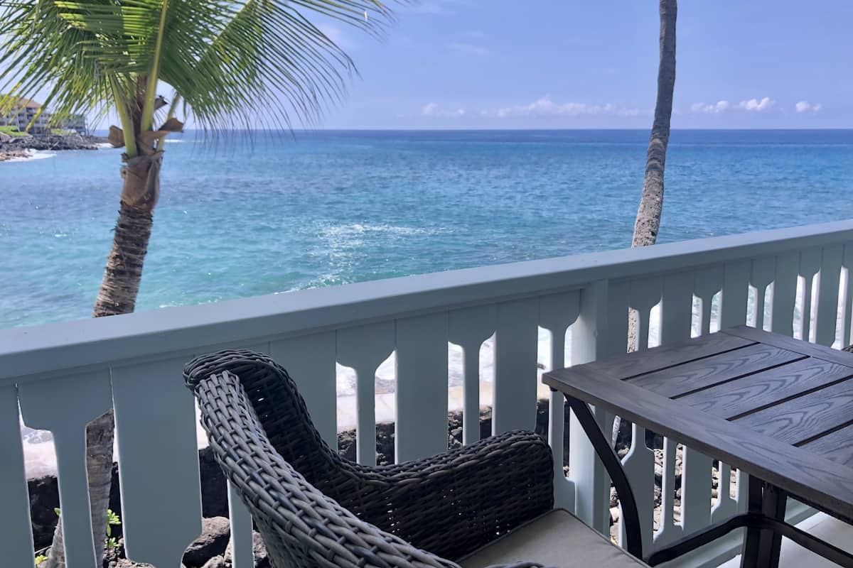 A balcony with wicker furniture overlooking a beach with clear blue water and palm trees.