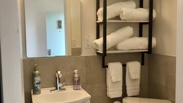 Un baño con lavamanos blanco, espejo, repisa con toallas, inodoro y un cubo de basura.