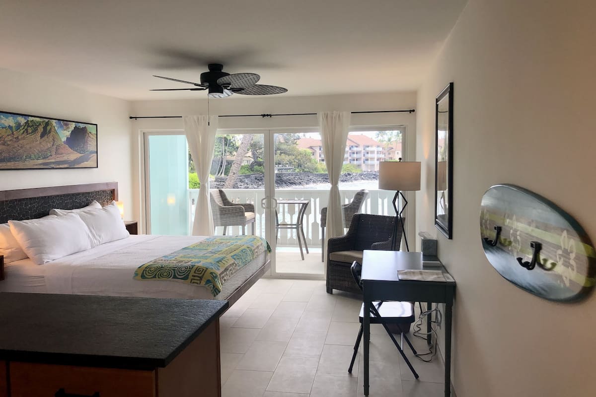 A hotel room with a bed, a desk, a chair, a ceiling fan, and a balcony with a view of the city.