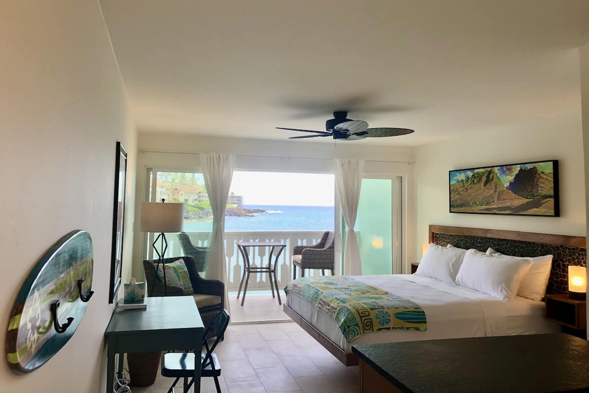 A hotel room with a bed, a desk, a chair, a balcony with a view of the ocean, and a ceiling fan.
