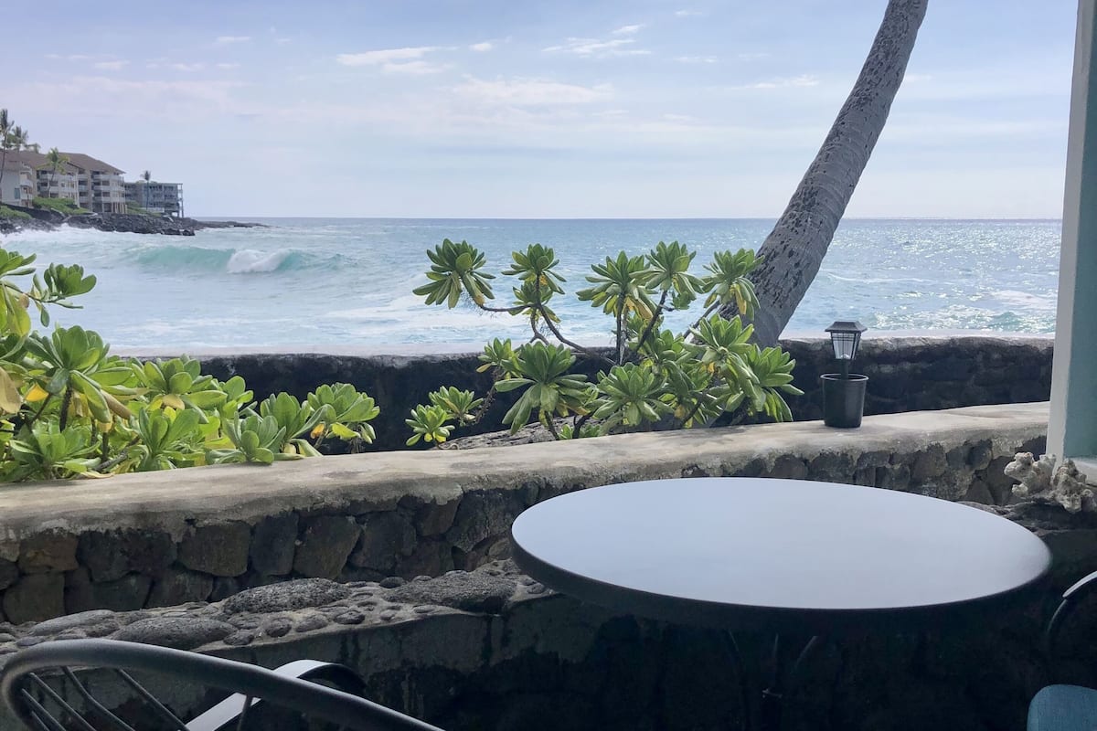 A patio with a round table and two chairs, overlooking the ocean with waves crashing onto a rocky shore.