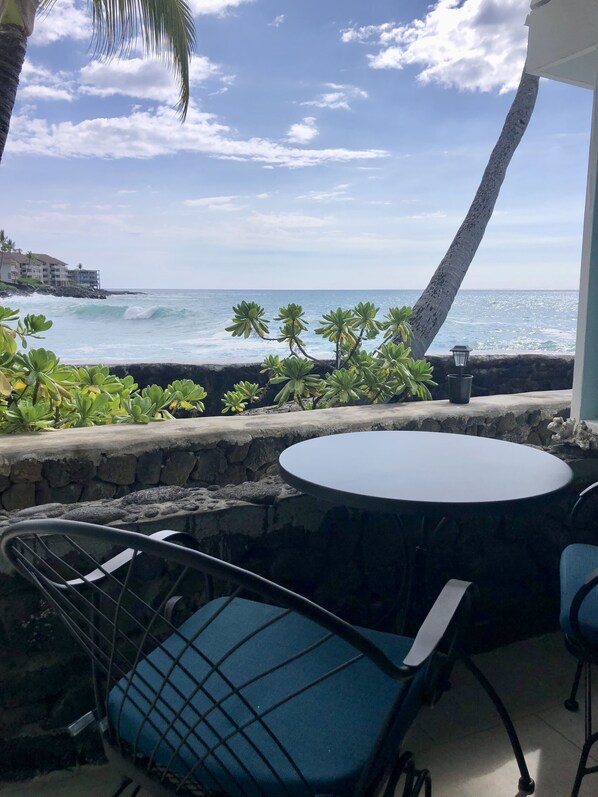 A patio with a round table and two chairs, overlooking the ocean with waves crashing onto a rocky shore.