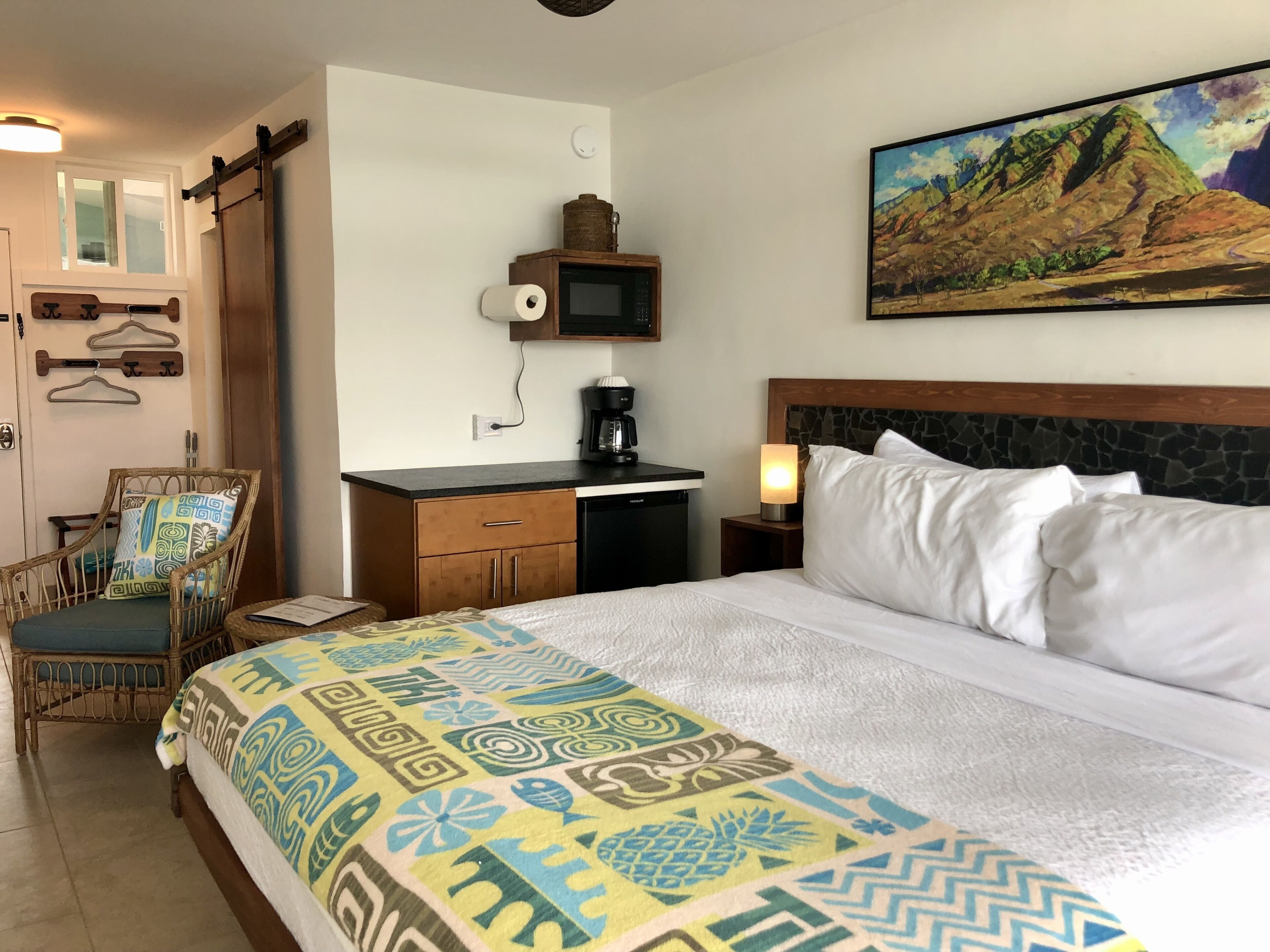 A hotel room with a bed, a desk with a coffee maker, a chair, a hanging coat rack, and a painting on the wall.