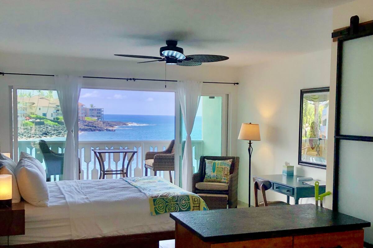 A hotel room with a bed, a desk, a chair, a ceiling fan, and a view of the ocean.
