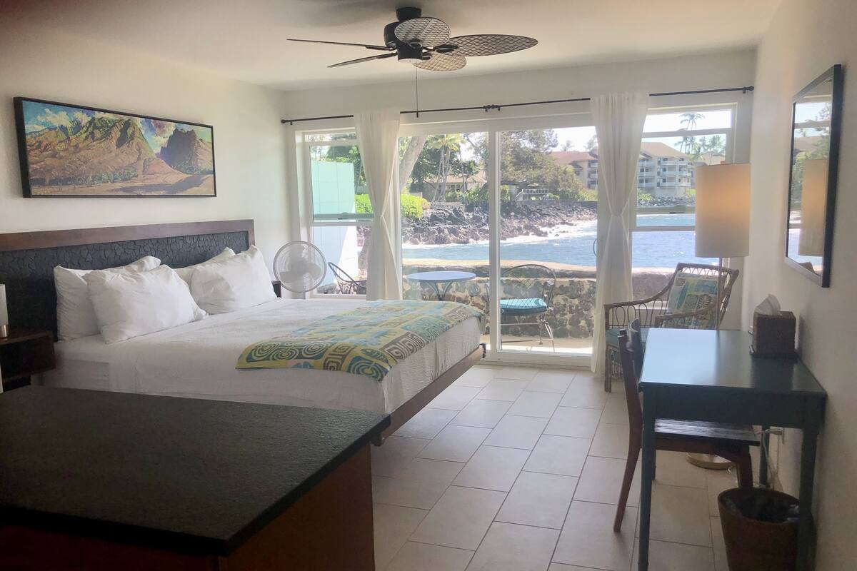 A hotel room with a bed, a desk, a chair, a fan, and a view of the ocean.