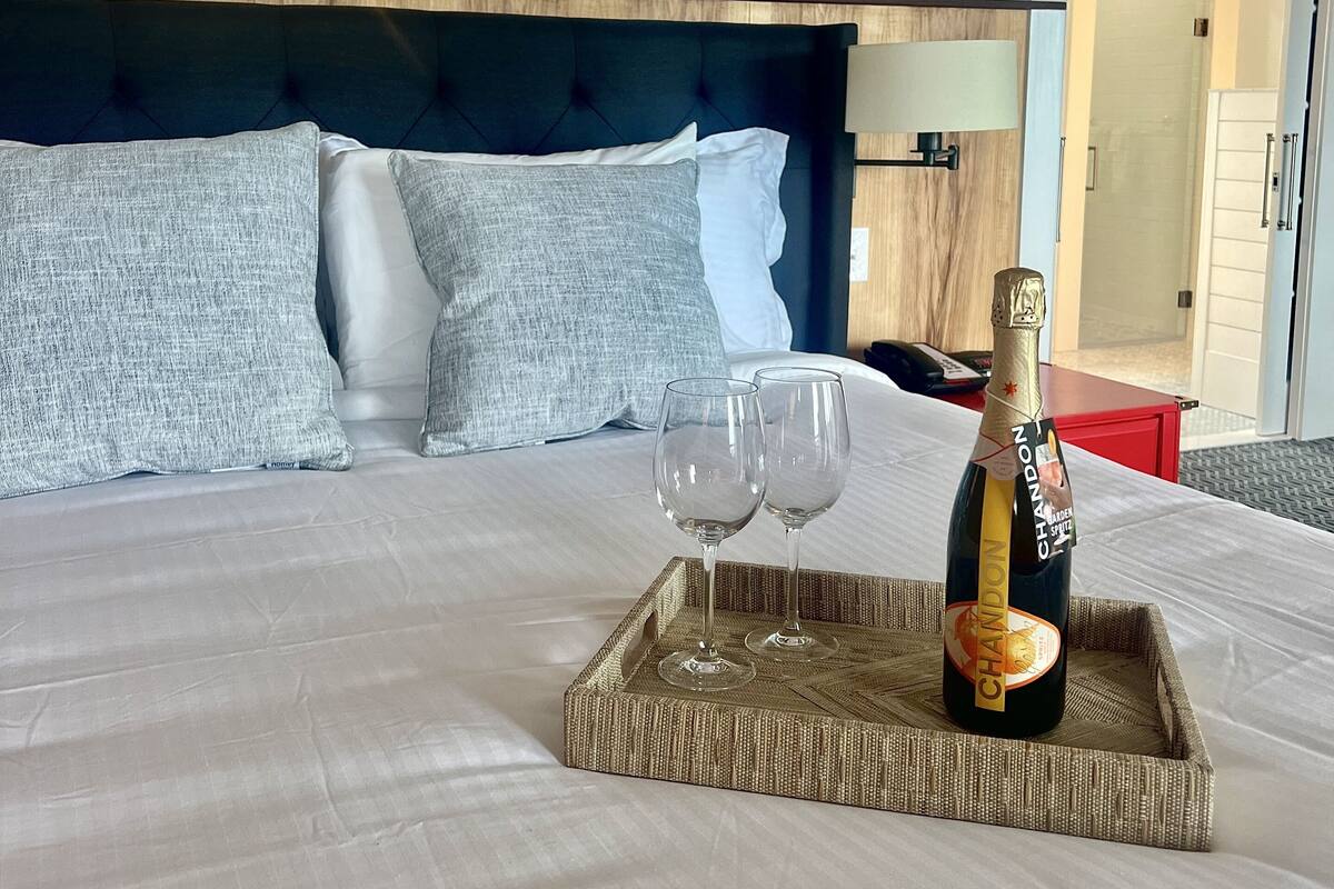 A neatly made bed with a tray holding a bottle of Chandon champagne and two glasses, a large headboard, and a modern chandelier.