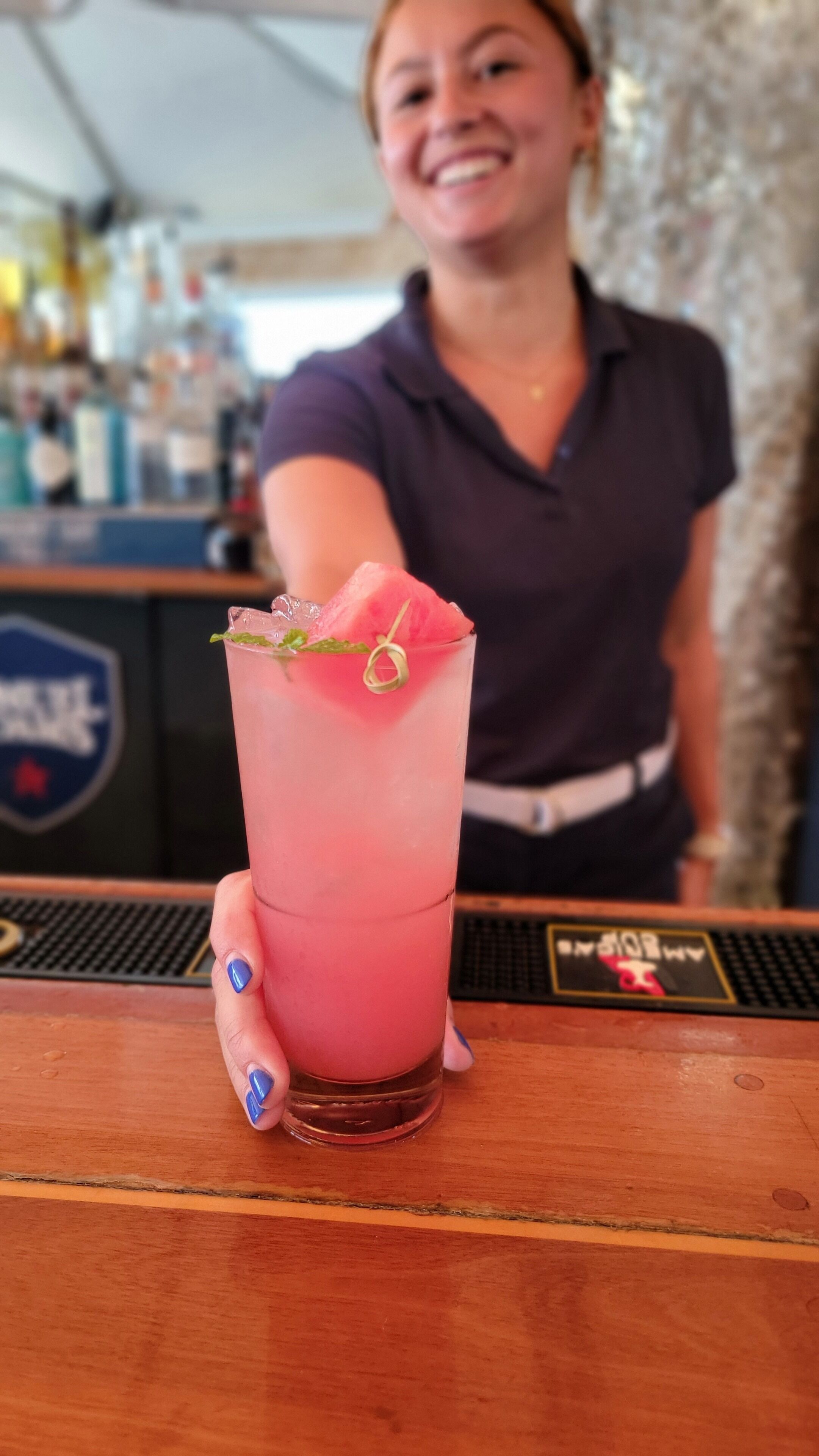 Un bar con una bebida en un vaso alto, una persona al fondo y un logotipo en la barra.