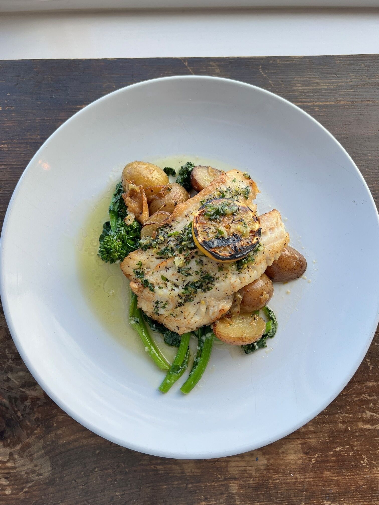 Une assiette blanche contenant un plat de poisson, des pommes de terre et des légumes verts, posée sur une table en bois.