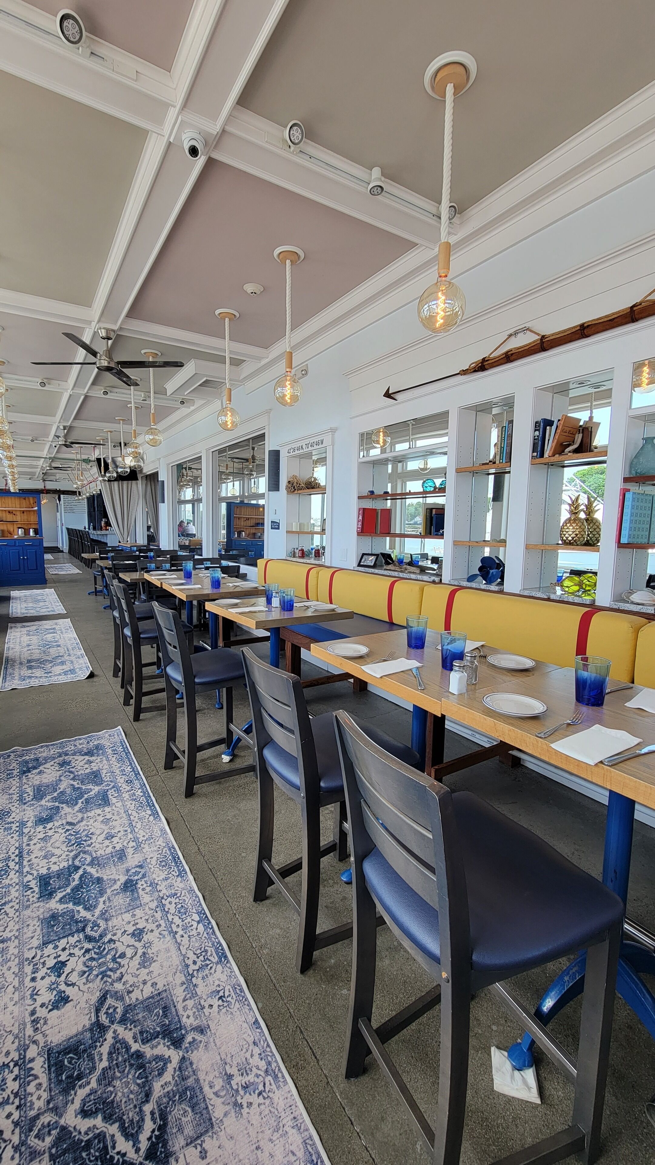 Ein Restaurant mit langen Gemeinschaftstischen, blau-weißen Teppichen mit Muster und einer gelben Sitzgruppe.