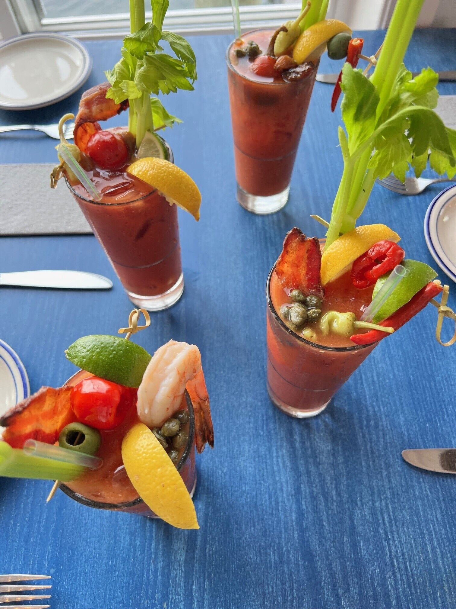 Drei Bloody Mary Cocktails, verziert mit verschiedenen Früchten und Gemüsesorten, serviert in hohen Gläsern auf einem blauen Tisch.