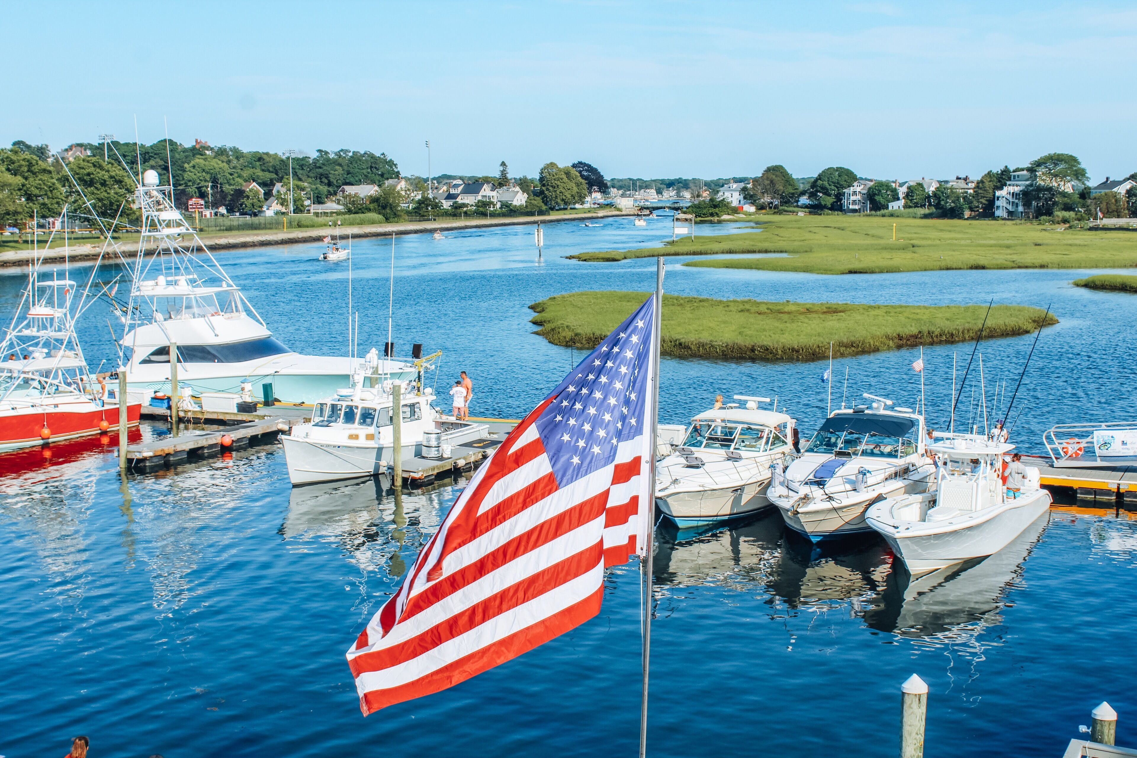 Una marina con diverse imbarcazioni ormeggiate, una bandiera americana ben visibile e una distesa d'acqua calma sullo sfondo, con vegetazione.