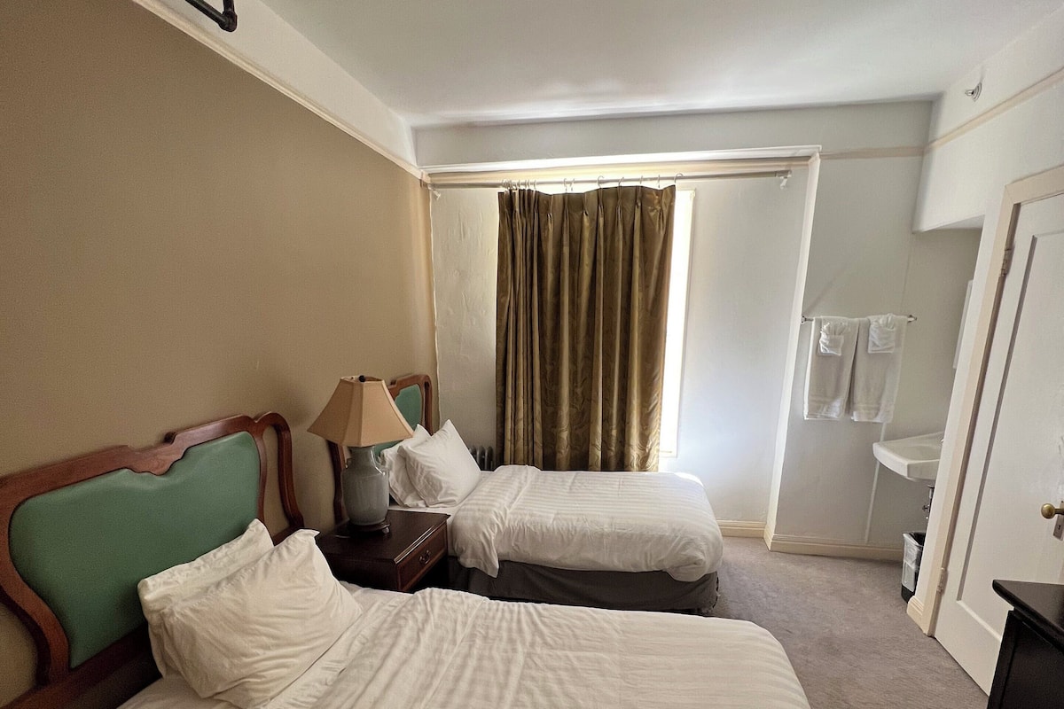 A hotel room with two beds, a nightstand, a lamp, a window with curtains, a door, and a ceiling fan.