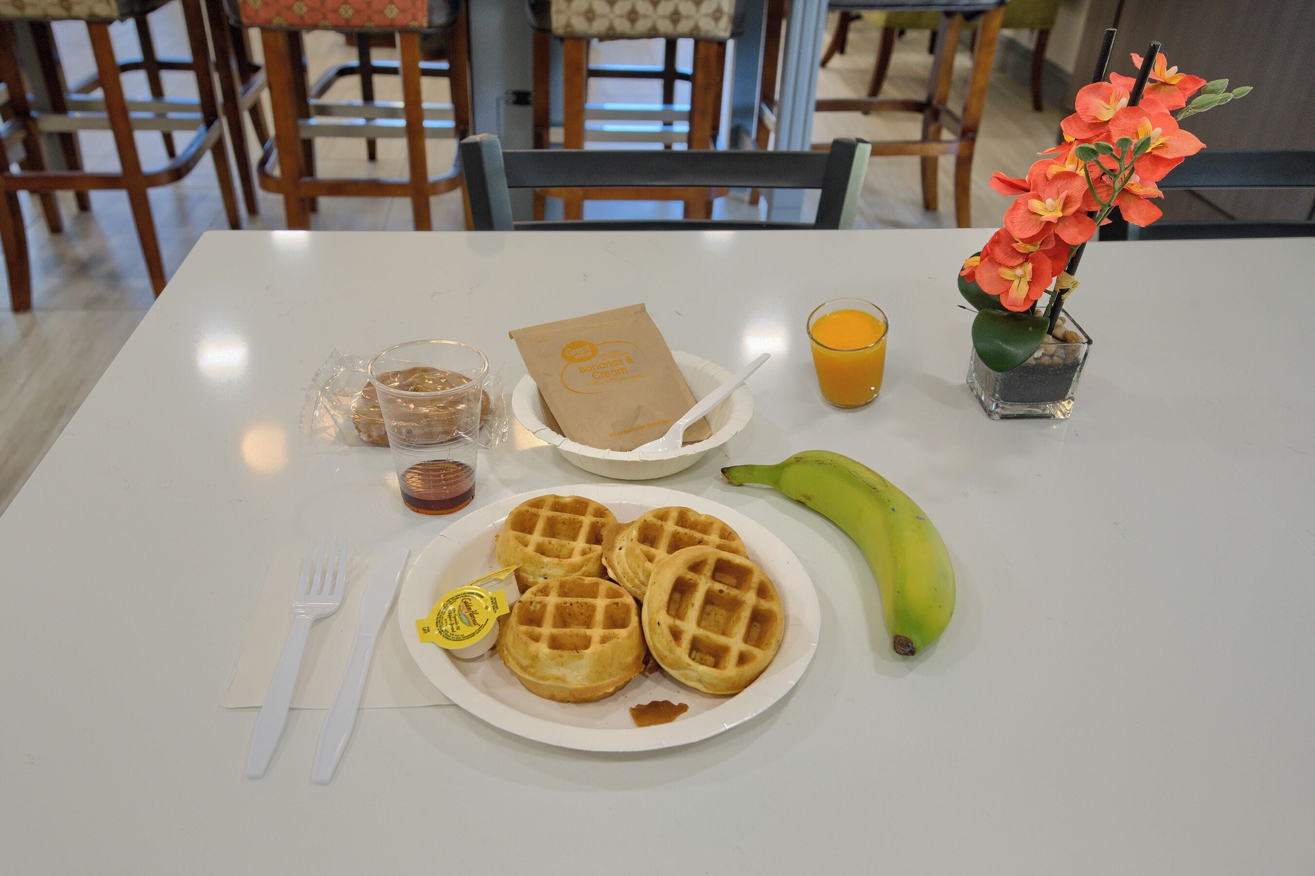 A breakfast table with waffles, a banana, a juice glass, a flower vase, and a coffee cup.