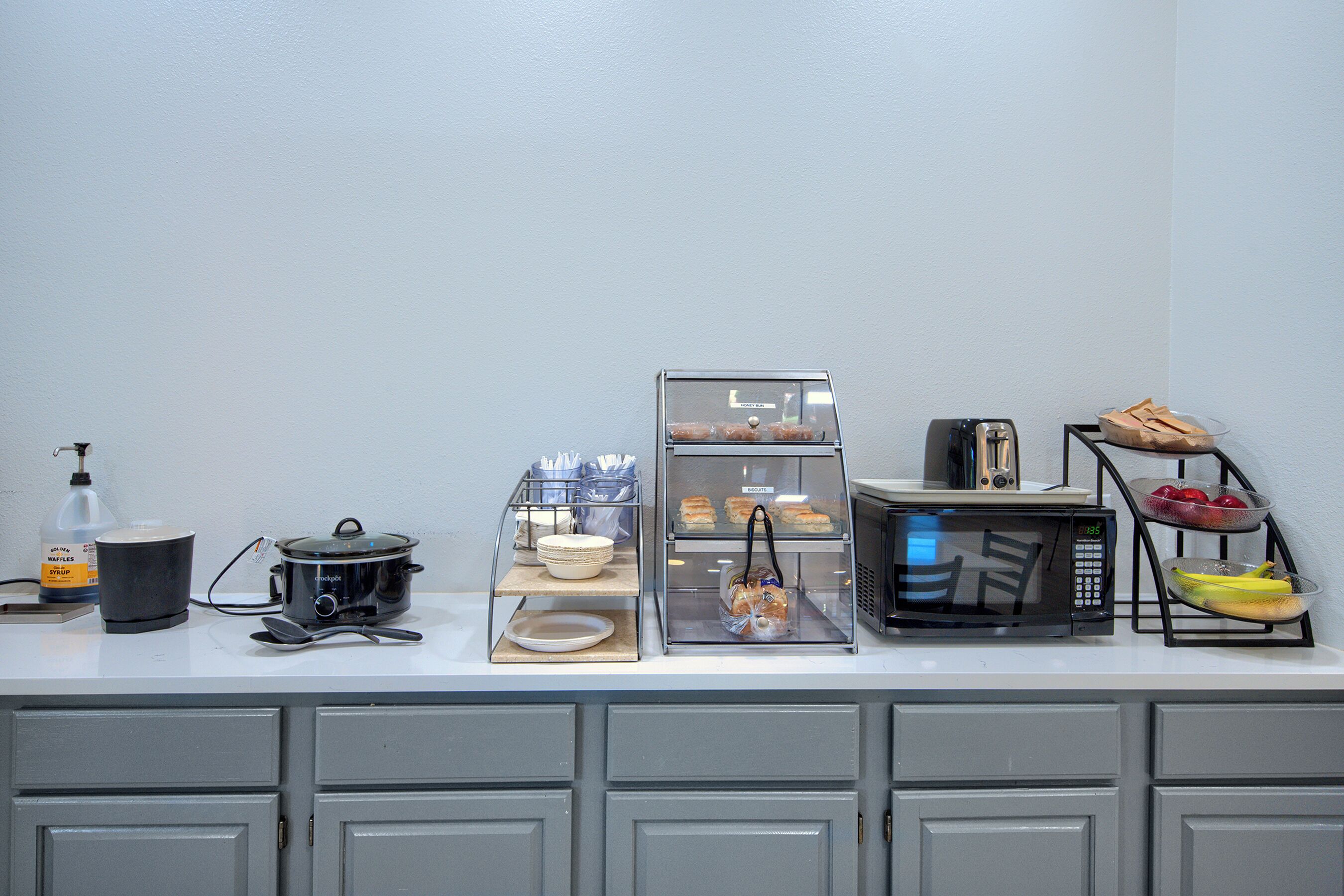 A kitchen counter with a slow cooker, a bread display, a microwave, and a fruit bowl.