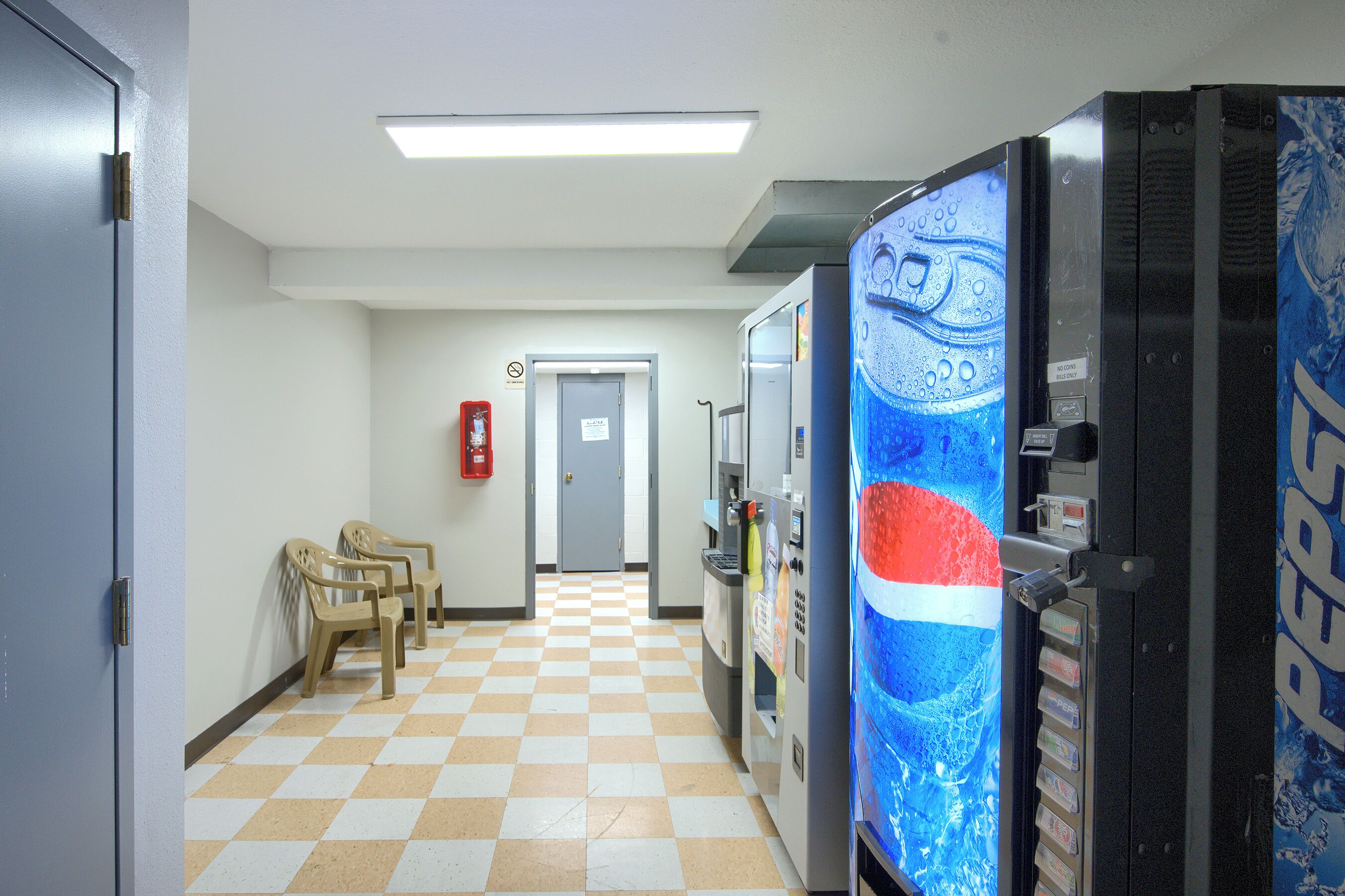 A hallway with a checkered floor, vending machines, and plastic chairs.