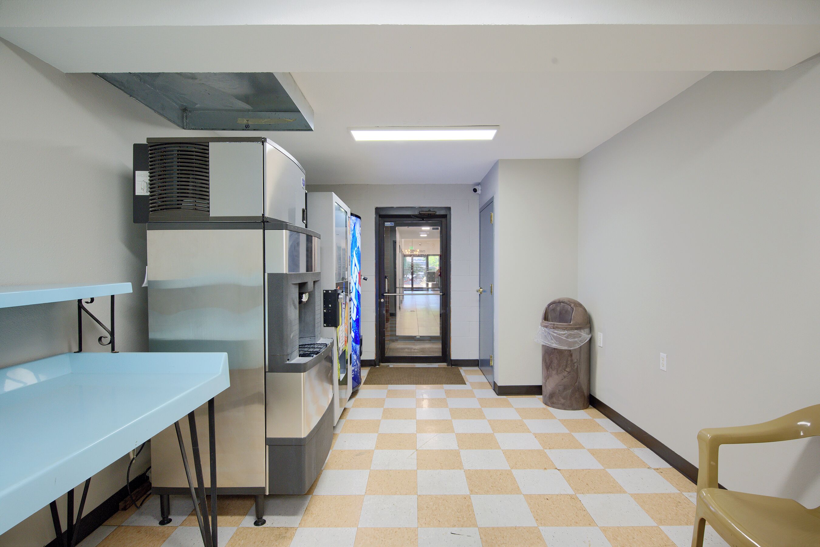A hallway with a checkered floor, a vending machine, a trash bin, and a blue table.