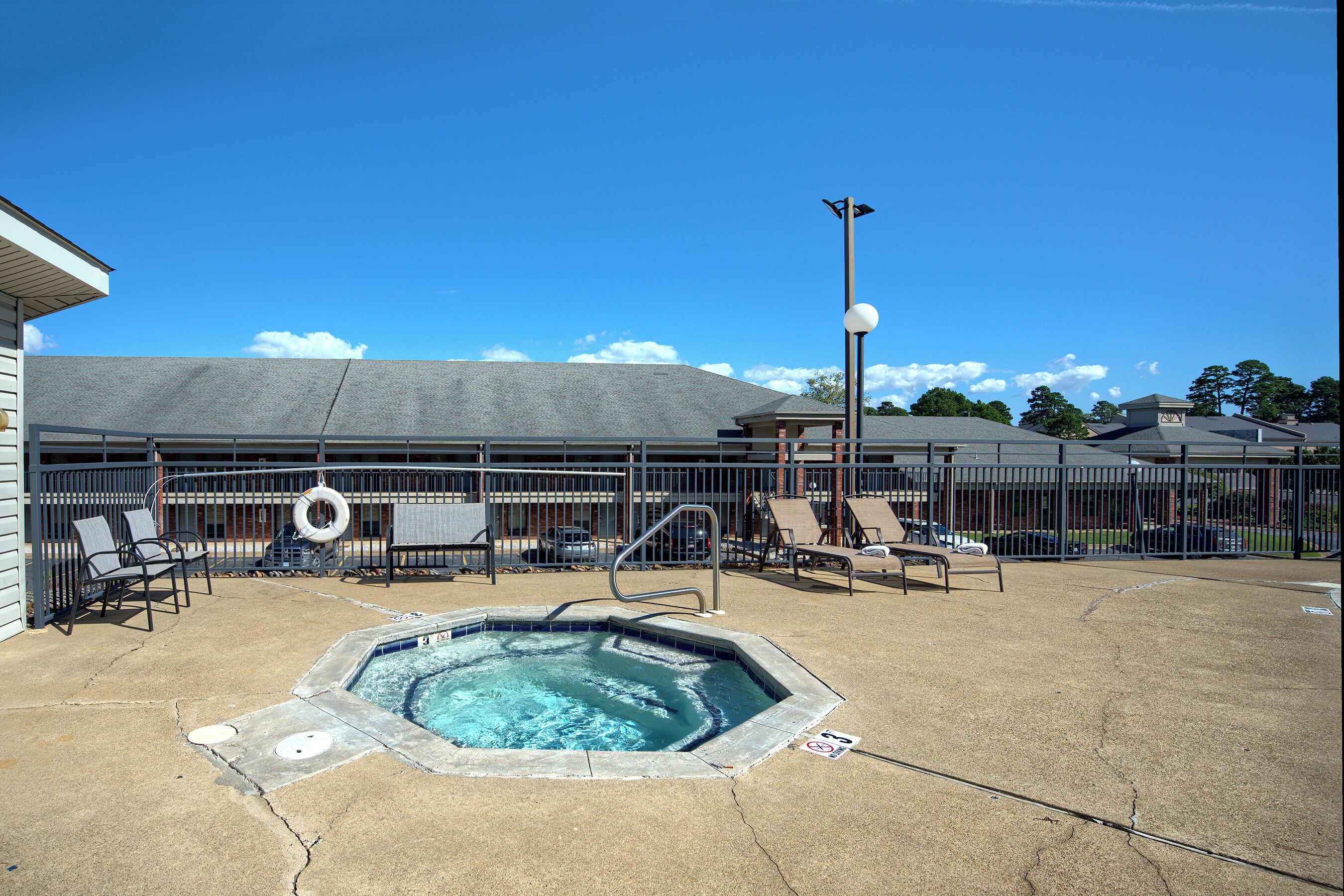 Une piscine dotée de chaises longues et d’un jacuzzi, entourée d’une clôture et bordée d’un bâtiment dont le toit est en pente.