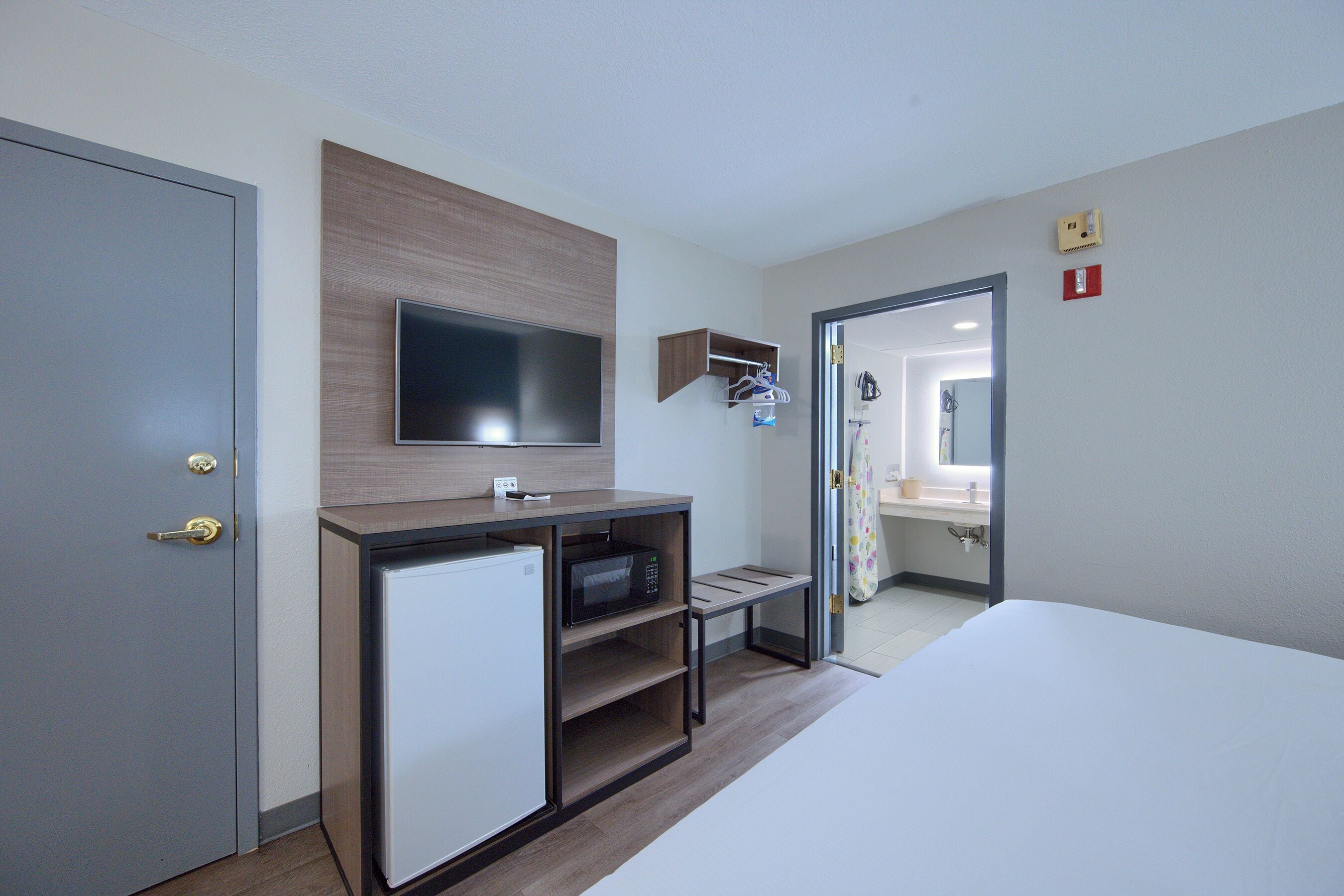 A hotel room with a bed, a television mounted on a wooden panel, a small refrigerator, a microwave, a shelf with hanging items, and a door leading to a bathroom.