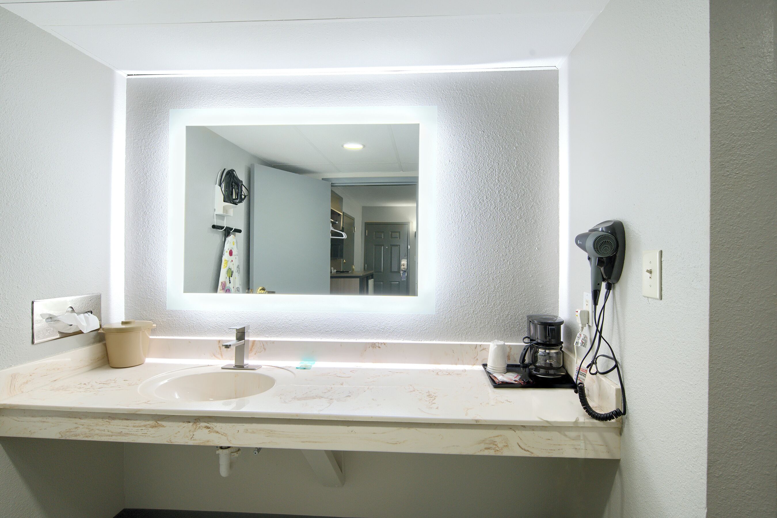 A bathroom with a large mirror, a sink, a hairdryer, and a coffee maker.