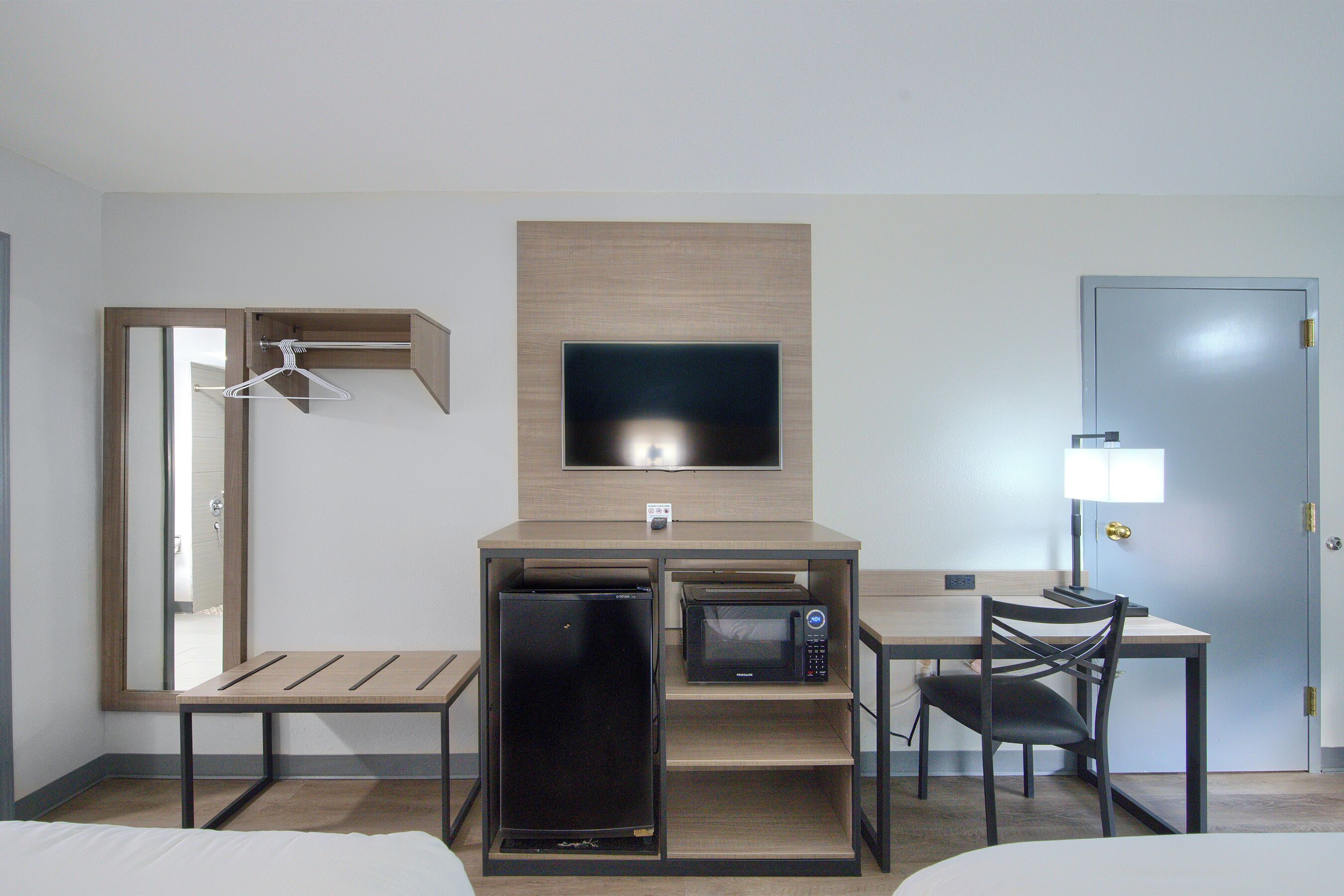 A compact hotel room with a bed, a desk with a computer, a microwave, a refrigerator, a TV, and a chair.