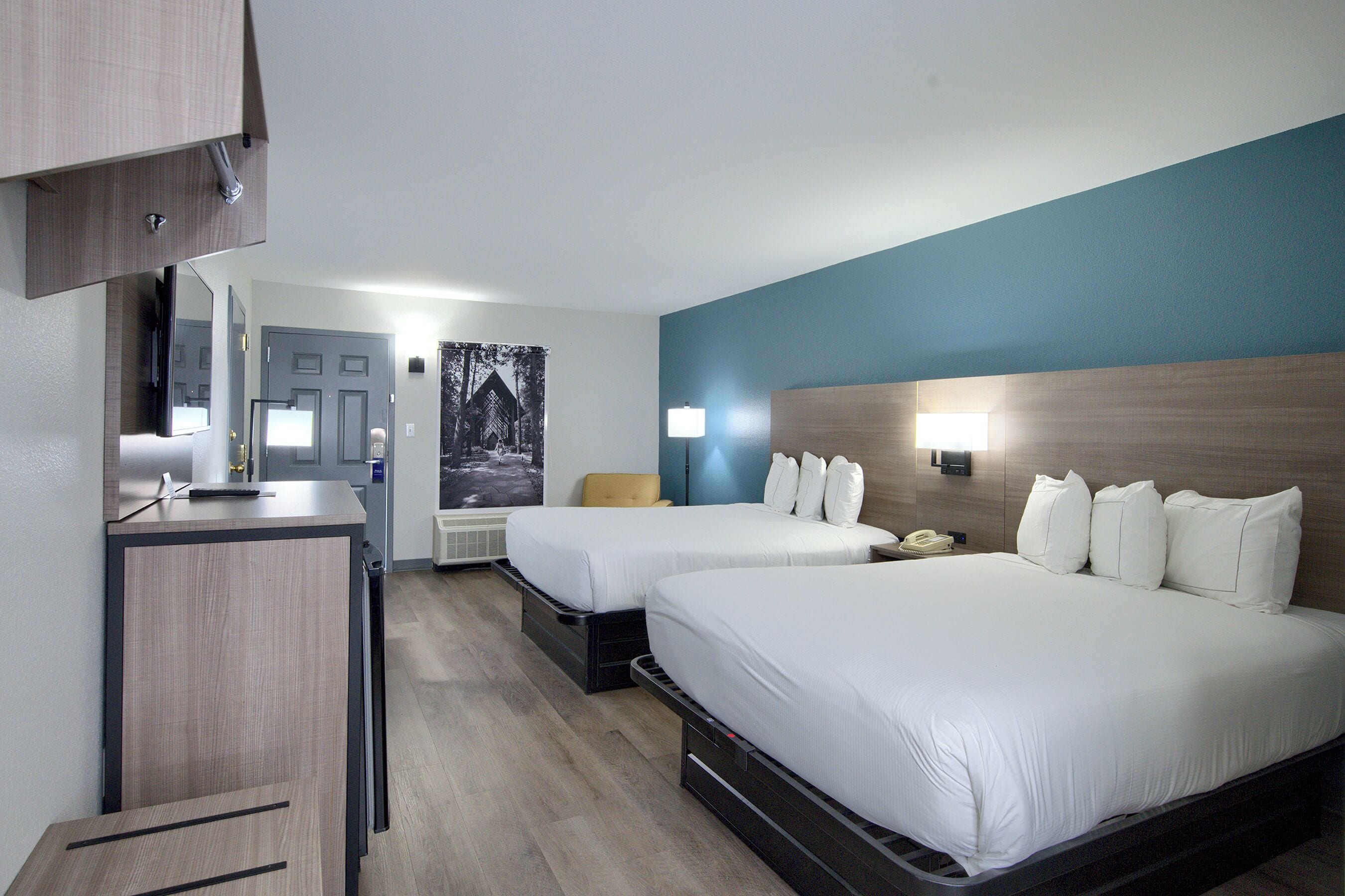 A hotel room with two beds, a desk, a chair, and a wall-mounted lamp.