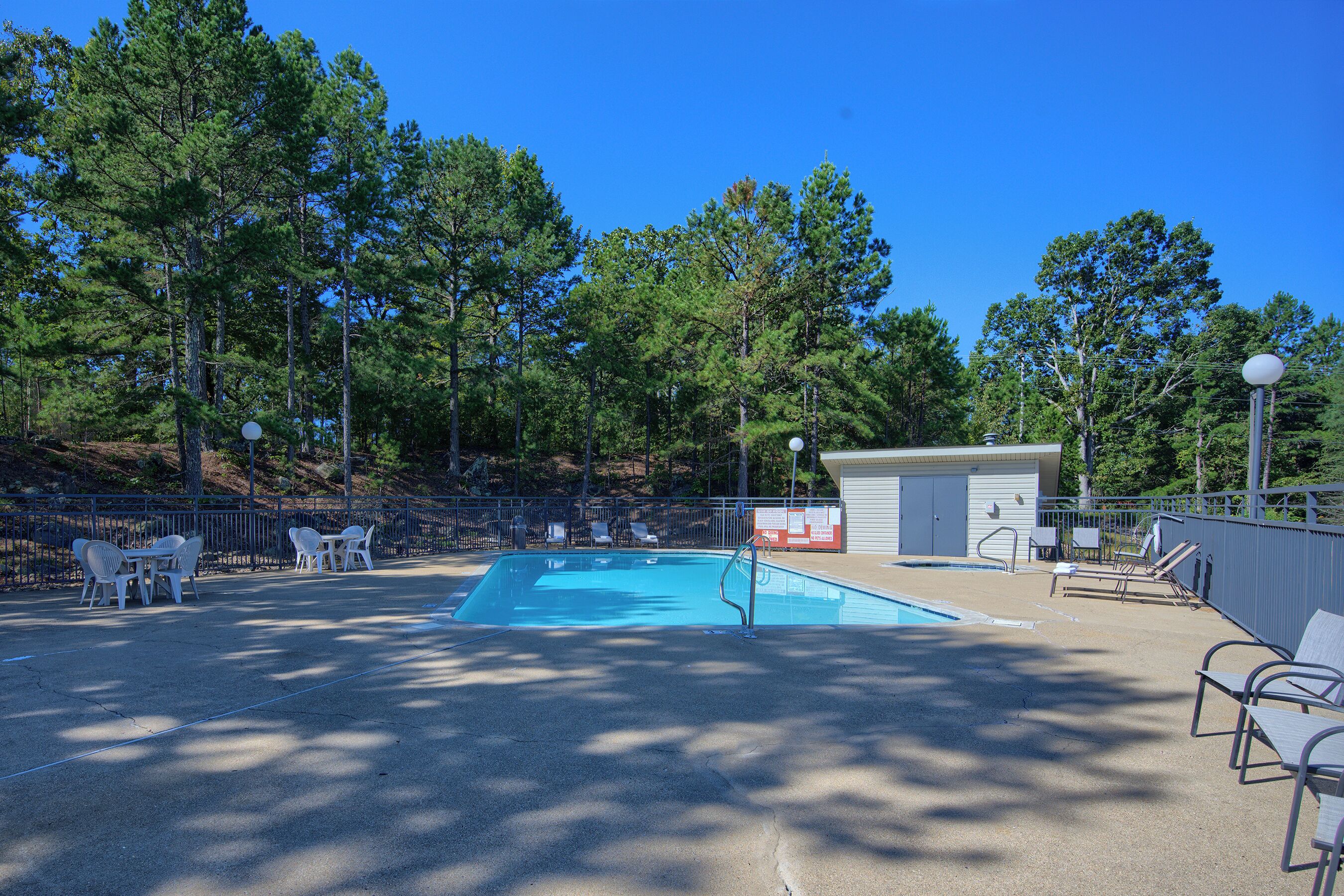 Piscina al aire libre con sillas y un pequeño edificio, rodeado de árboles.