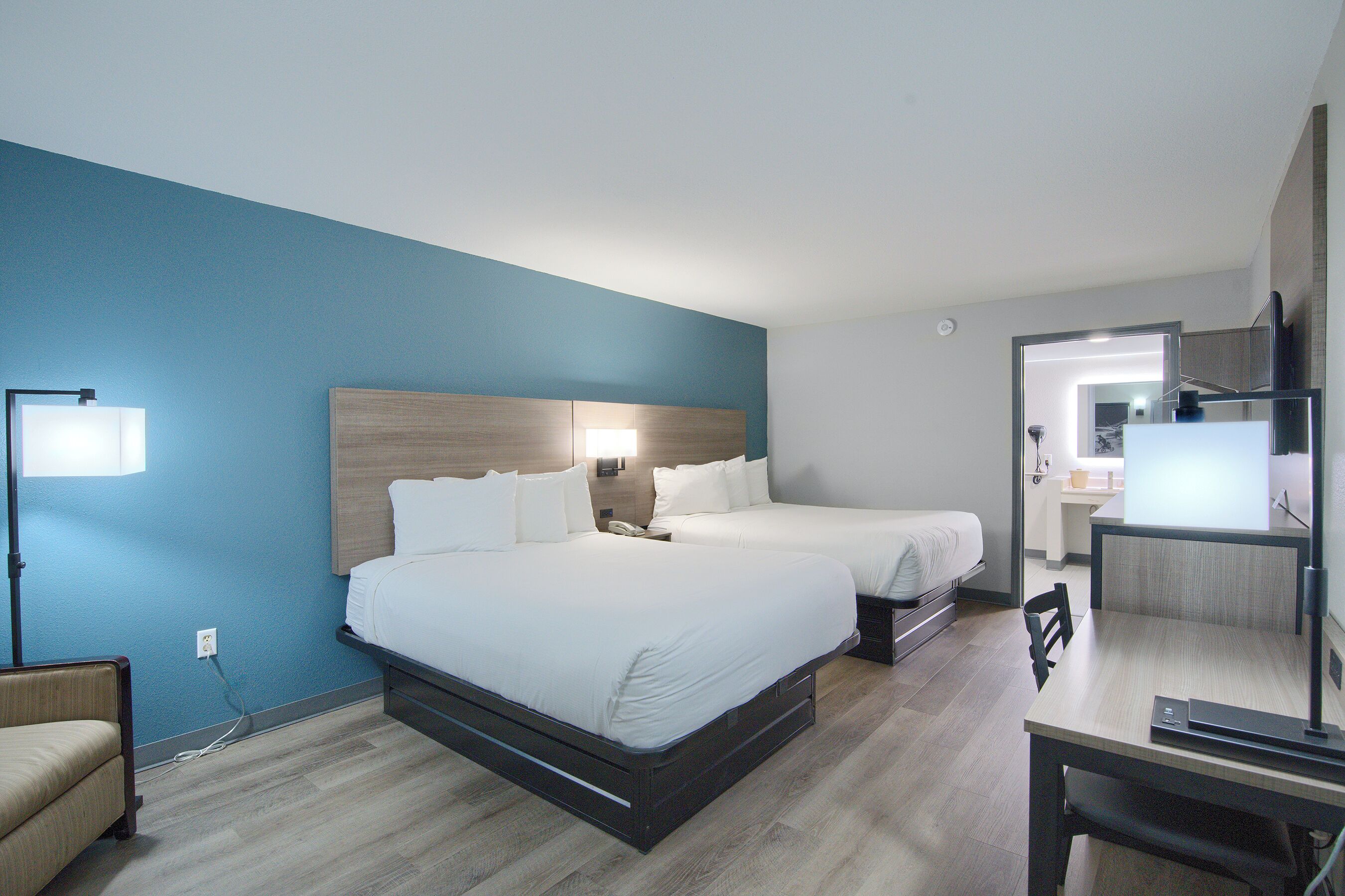 A hotel room with two beds, a desk, a chair, a lamp, and a TV.