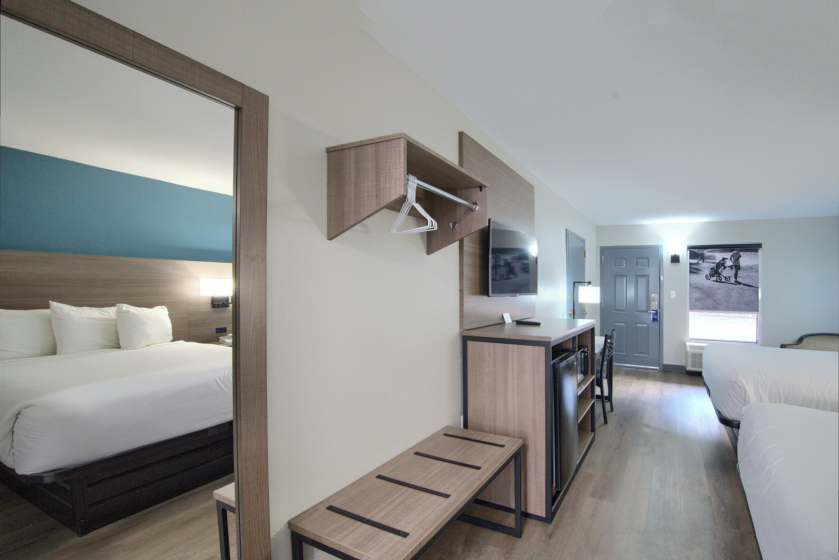 A hotel room with a bed, a desk, a chair, a bench, a TV, and a door.