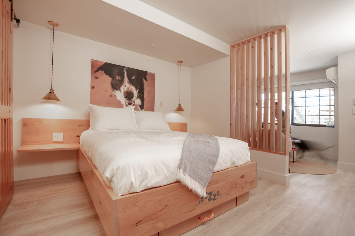 A bedroom with a wooden bed, white bedding, a dog-themed wall art, and a desk with a chair.