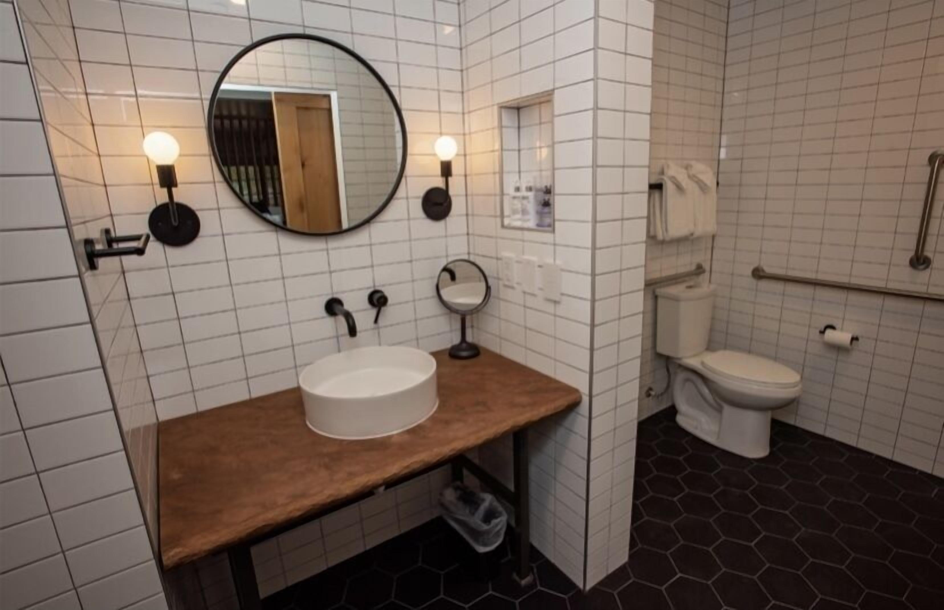 Un bany modern amb un mirall rodó, un tocador de fusta, un lavabo blanc, un vàter i una barana.