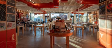 Een ruim restaurant met hoge plafonds, zichtbare leidingen en grote ramen. Tafels staan klaar voor dineren, en er liggen diverse gerechten en decoraties op de tafels.