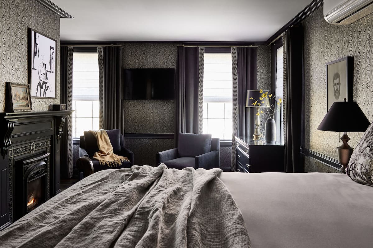 A hotel room with a fireplace, a bed, a chair, a TV, and a window with curtains.