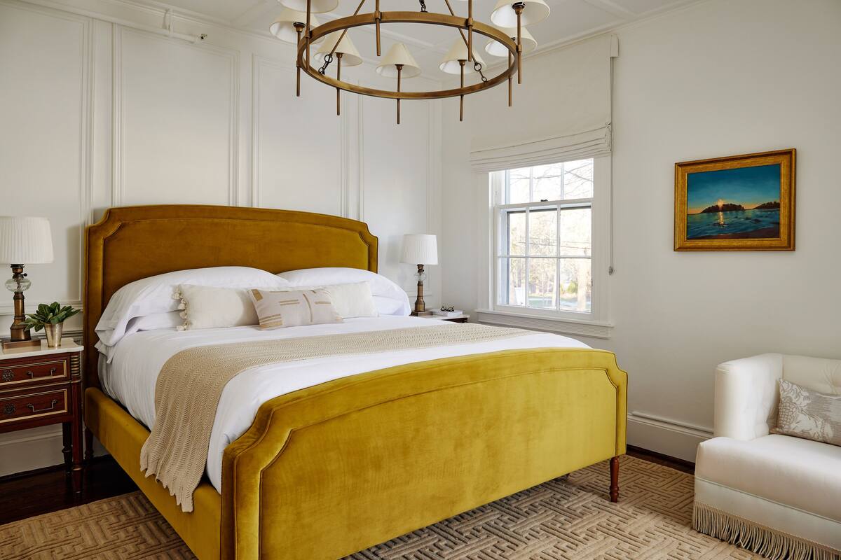 A neatly arranged bedroom with a large bed, bedside lamps, a window with a view, and a painting on the wall.