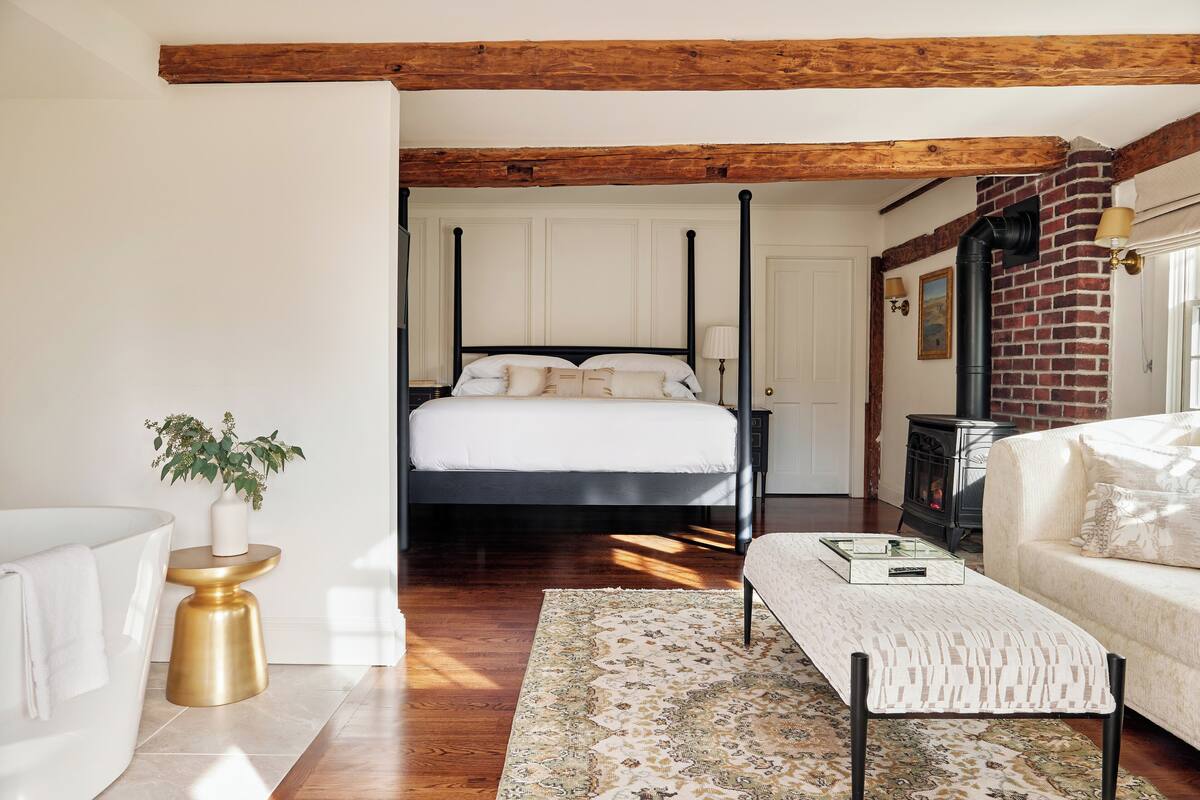A bedroom with a four-poster bed, a white sofa, a patterned rug, and a small side table with a plant.