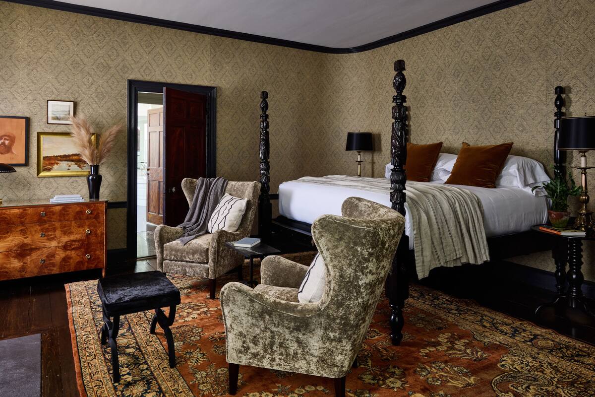 A bedroom with a four-poster bed, a wooden dresser, two armchairs, and a patterned rug.