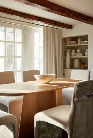 A dining area with a wooden table, chairs, and a large bowl. There is a window with curtains and a shelf with decorative items. - Kennebunkport Captains Collection (Kennebunkport)