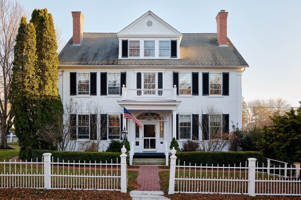 A two-story white house with a front porch, a brick chimney, and a white picket fence. - Kennebunkport Captains Collection (Kennebunkport)