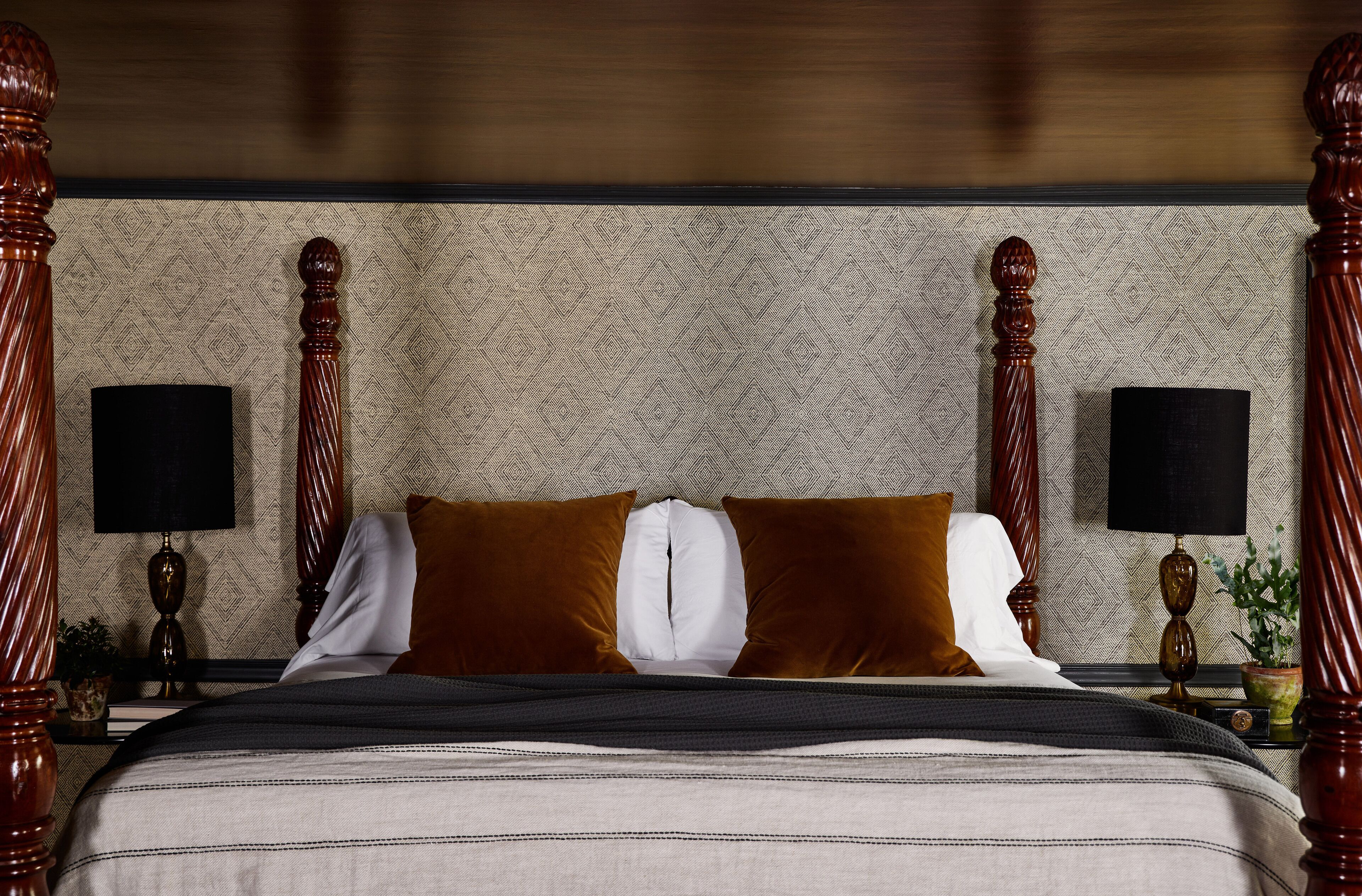 Una cama bien hecha con una colcha a rayas, dos almohadas marrones y una blanca. Hay dos mesitas de noche con lámparas y una planta en maceta.